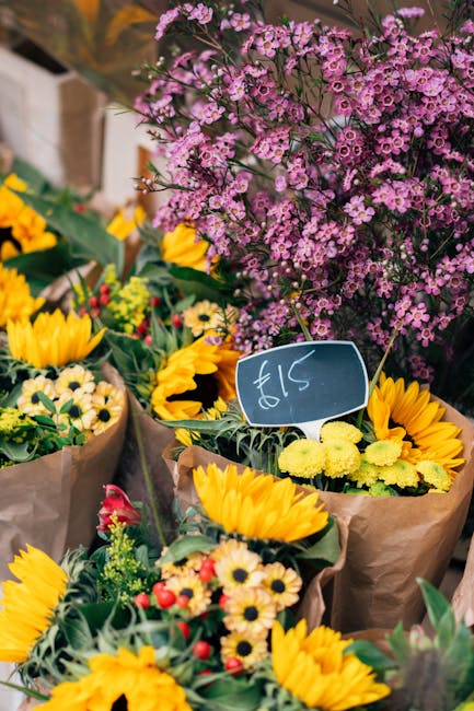 A vibrant display of fresh floral bouquets featuring bright yellow sunflowers, yellow and white daisies, and small red and yellow flowers, wrapped in brown kraft paper and arranged outdoors. In the ba