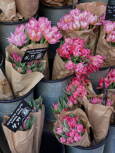 An arrangement of fresh pink tulips displayed in metallic buckets, each wrapped in brown paper with handwritten chalkboard labels showing prices and plant types. The tulips vary in shades from soft pa