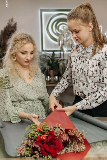Two women are showcased in a floral shop preparing a bouquet arrangement. The woman on the right, with long, light brown hair tied in a high ponytail, is wearing a patterned blouse with a floral and g